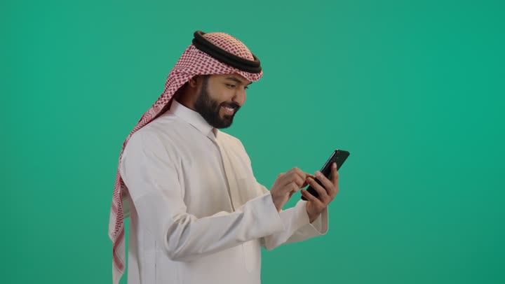 استخدام الأجهزة التقنية الحديثة و المتطورة, تصفح وسائل التواصل الاجتماعي عبر الجوال, رجل عربي خليجي سعودي مبتسم يرتدي الثوب التقليدي و الشماغ يمسك الهاتف المحمول بيده، خلفية خضراء
