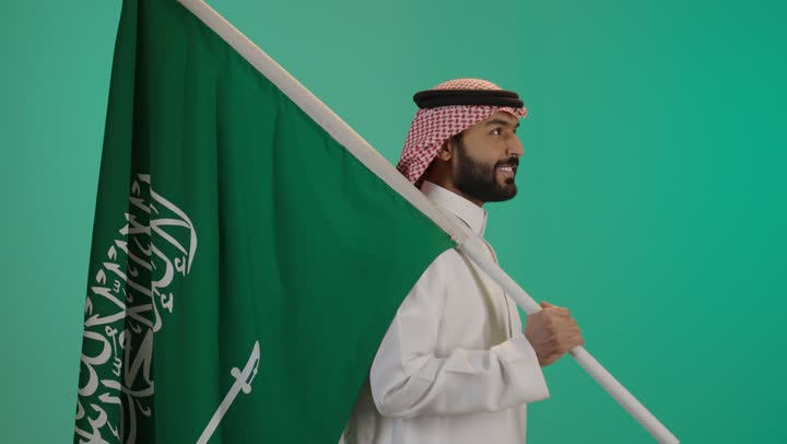 Commemorating the Saudi National Day, a portrait of a young Arab Gulf Saudi man wearing traditional attire and a shemagh, carrying the Kingdom's flag on his shoulder with gestures of pride, participating in the celebration of national and popular occasions and holidays, with a green background.