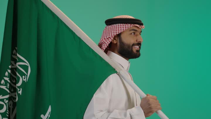 بورتريه لشاب عربي خليجي سعودي يرتدي الثوب التقليدي والشماغ يحمل علم المملكة على كتفه بإيماءات الاعتزاز، إحياء ذكرى اليوم الوطني السعودي، المشاركة في الاحتفال بالمناسبات والأعياد الوطنية والشعبية، خلفية خضراء