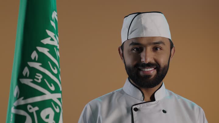بورتريه لشيف عربي خليجي سعودي يرتدي الزي الرسمي الخاص بمزاولة مهنة الطباخ بمسك علم المملكة، تحقيق النمو والتطوربسواعد أبناء الوطن, الإحتفال بذكرى اليوم الوطني السعودي 23 سبتمبر والعيد الوطني، الاخلاص والولاء للبلاد، خلفية بنية