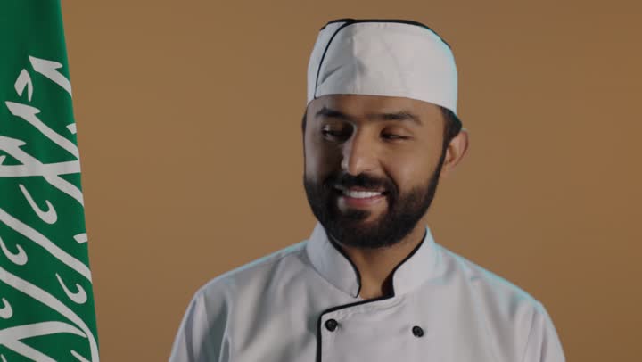 احتراف مهنة وفنون الطبخ، بورتريه لشيف عربي خليجي سعودي يرتدي الزي الرسمي الخاص بمزاولة مهنة الطباخ بمسك علم المملكة و ينظر اليه بايماءات الفخر, الإحتفال بذكرى اليوم الوطني السعودي 23 سبتمبروالعيد الوطني، خلفية بنية