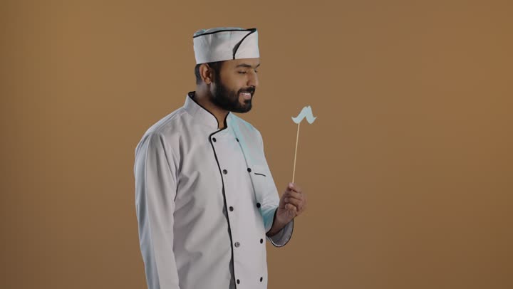 بورتريه لرجل عربى  خليجي سعودى يرتدي الزي الرسمي الخاص بمزاولة مهنة الطباخ يمسك بيده عود خشبي مع قصاصة ورقية على شكل شارب، الاحتفال السنوي باليوم العالمي للرجال ، ١٩ نوفمبر  ، الاهتمام بصحة الرجل، خلفية بنية