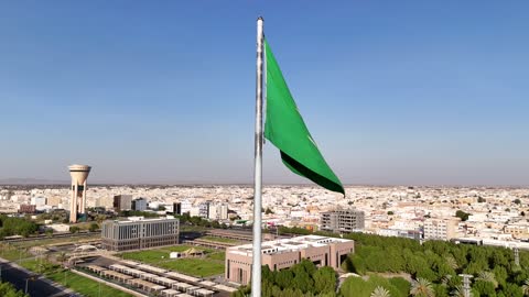 The tallest flagpole in the world is in Tabuk, Saudi National Day, drone footage from above of the largest flagpole in the city of Tabuk in the Kingdom of Saudi Arabia, traffic movement, roads and streets of Tabuk, famous tourist landmarks of Tabuk, the model of the official emblem of the Kingdom of Saudi Arabia in Tabuk Square.