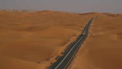 The sand hills and rock formations in the Arab deserts, recreational activities for driving cars in desert areas, the sand dunes in the Kingdom of Saudi Arabia, drone footage of a Mercedes on an asphalt road between the desert and the golden sand dunes.