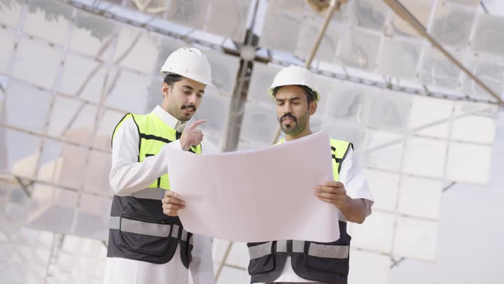 تبادل الخبرة و الرأى, مهندسان عربيان خليجيان سعوديان يحملان مخطط هندسى و يشرفان على محطات الطاقة الشمسية ينظران الى شىء ما ، إرتداء خوذة وسترة الحماية ، الإشراف على العمل، أنظمة الطاقة الشمسية
