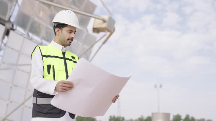 Review of the engineering plan, supervision of the engineering project, a Saudi Gulf Arab engineer holding an engineering design, wearing a helmet and safety vest, Saudi professions and jobs, solar power stations.
