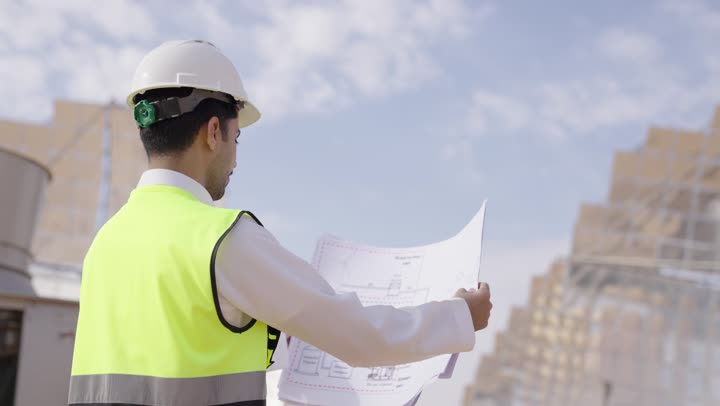تصوير من الخلف لمهندس عربى خليجى سعودى يمسك مخطط و تصميم هندسى ينظر الى شىء ما, إرتداء خوذة وسترة الحماية, مهن ووظائف سعودية، محطات الطاقة الشمسية