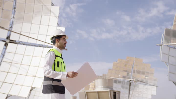 دراسة المخطط والمشروع على ارض الواقع، مهنة ووظيفة هندسية, إرتداء خوذة وسترة الحماية, تصوير من الجانب لمهندس عربى خليجى سعودى يمسك مخطط و تصميم همدسى يمشى فى الموفع