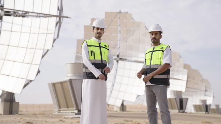 النظر الى الكاميرا بتعابير السرور و الثقة, الوقوف بأيدى متقاطعة, إرتداء خوذة وسترة الحماية, مهندسان عربيان خليجيان سعوديان يشرفان على محطات الطاقة الشمسية
