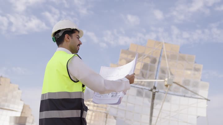 Wearing a helmet and protective vest, a side and back view of a Saudi Gulf Arab engineer holding a plan and engineering design looking at something, professions and jobs in Saudi Arabia, solar power stations.