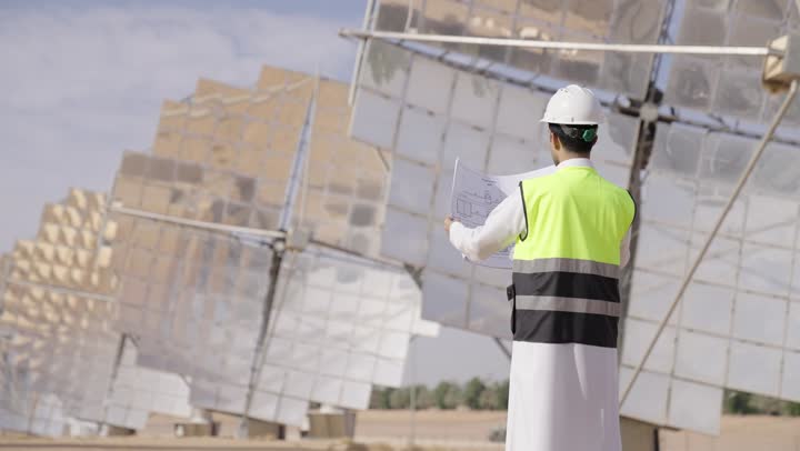 تصوير من الجانب و الخلف لمهندس عربى خليجى سعودى  يمسك مخطط و تصميم هندسى ينظر الى شىء ما, إرتداء خوذة وسترة الحماية, مهن ووظائف سعودية، محطات الطاقة الشمسية