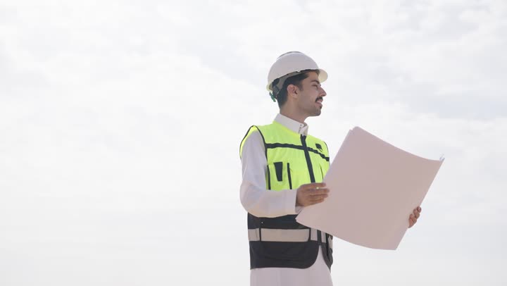 مفهوم هندسة الطاقة, تصوير مقرب لمهندس عربى خليجى سعودى  يمسك مخطط و تصميم هندسى ينظر الى شىء ما, إرتداء خوذة وسترة الحماية, مهن ووظائف سعودية، محطات الطاقة الشمسية