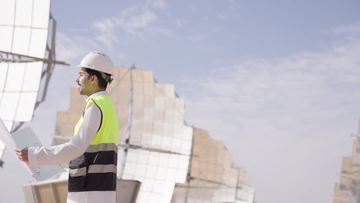 Auditing and supervising work, a Saudi Gulf Arab engineer holding a plan and engineering design looking at something, wearing a helmet and safety vest, Saudi professions and jobs, solar power stations.