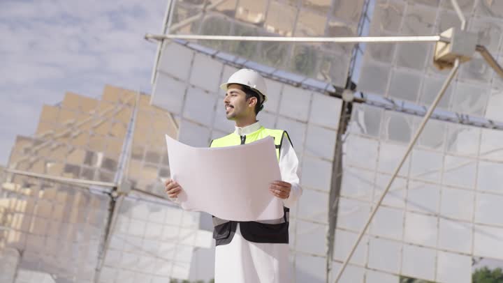 مهندس عربى خليجى سعودى  يمسك مخطط و تصميم هندسى ينظر الى شىء ما, التدقيق والإشراف على العمل, إرتداء خوذة وسترة الحماية, مهن ووظائف سعودية، محطات الطاقة الشمسية
