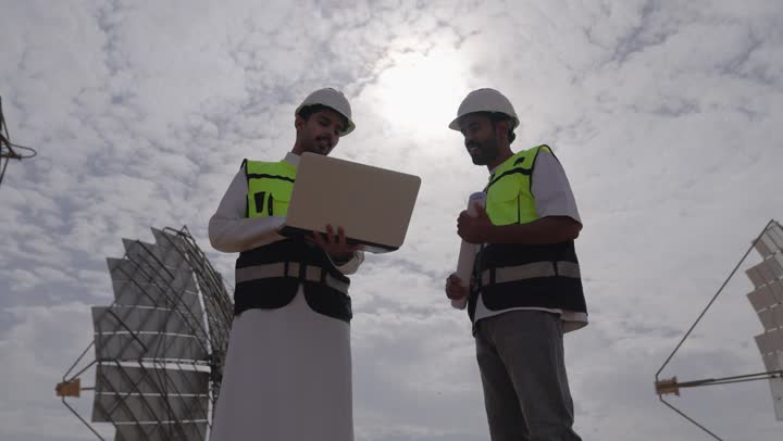 A bottom view of two Arab Gulf engineers from Saudi Arabia using a laptop overseeing solar power stations, wearing helmets and safety vests, supervising the work, solar power systems, exchanging expertise and opinions among colleagues.