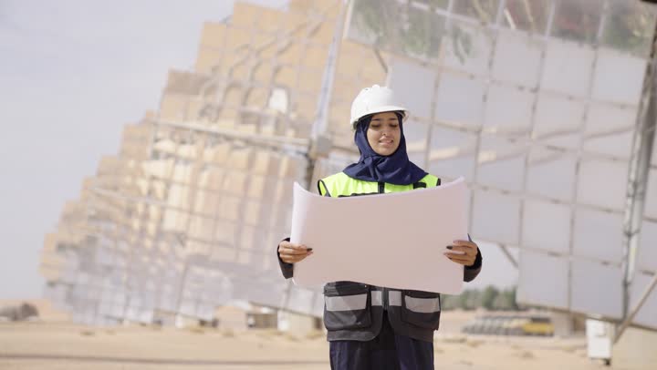 دراسة المخطط والمشروع على ارض الواقع, مهندسة عربية خليجية سعودية محجبة مبتسمة ترتدي العباءة السوداء و الخوذة وسترة الحماية تحمل تصميم هندسى, مهن ووظائف سعودية، محطات الطاقة الشمسي