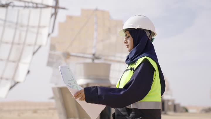 The concept of energy engineering, a side view of a smiling Saudi Gulf Arab female engineer wearing a black abaya, helmet, and safety vest, holding a plan and engineering design, professions and jobs in Saudi Arabia, solar power stations.