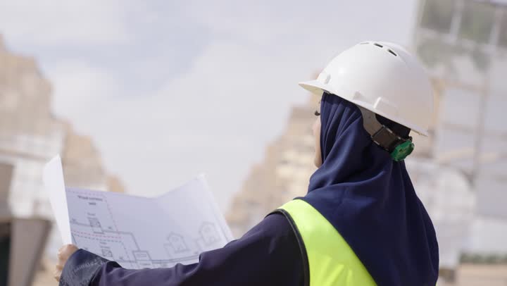 تصوير من الجانب لمهندسة عربية خليجية سعودية محجبة مبتسمة ترتدي العباءة السوداء و الخوذة وسترة الحماية تحمل مخطط و تصميم هندسى, مستقبل الطاقة المتجددة, مهن ووظائف سعودية، محطات الطاقة الشمسية