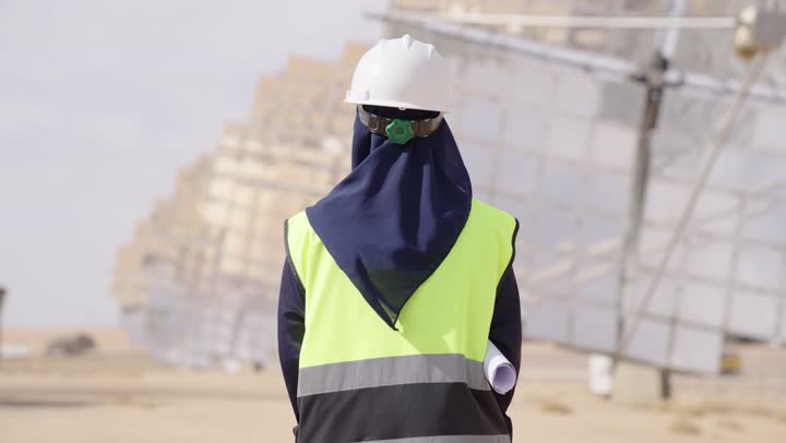 Auditing and supervising work, a rear view of a smiling Saudi Gulf Arab female engineer wearing a black abaya, helmet, and safety vest holding an engineering plan, the future of renewable energy, Saudi professions and jobs, solar power stations.
