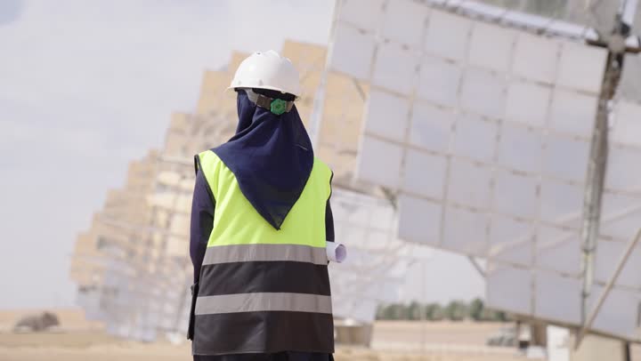 A rear view of a smiling Saudi Arabian Gulf female engineer wearing a black abaya, helmet, and safety vest, holding an engineering plan while supervising the project on site, the future of renewable energy, Saudi professions and jobs, solar power plants.