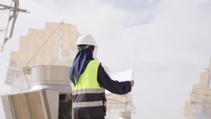 مهندسة عربية خليجية سعودية محجبة مبتسمة ترتدي العباءة السوداء و الخوذة وسترة الحماية تمسك بيدها مخطط المشروغ الهندسى, التدقيق والإشراف على العمل, مهن ووظائف سعودية,  محطات الطاقة الشمسية