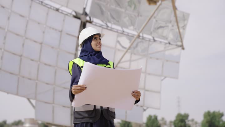 التدقيق والإشراف على العمل, مهن ووظائف سعودية, مهندسة عربية خليجية سعودية محجبة مبتسمة ترتدي العباءة السوداء و الخوذة وسترة الحماية تمسك بيدها مخطط المشروغ الهندسى تنظر الى شىء ما, محطات الطاقة الشمسية