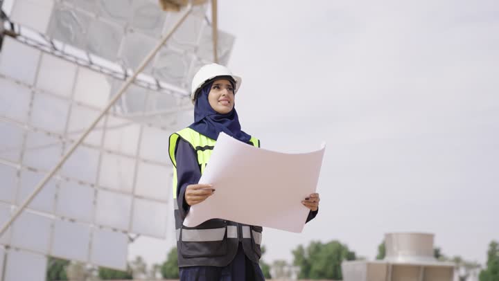 دراسة المخطط والمشروع على ارض الواقع, مهندسة عربية خليجية سعودية محجبة مبتسمة ترتدي العباءة السوداء و الخوذة وسترة الحماية تمسك بيدها مخطط المشروغ الهندسى تنظر الى شىء ما, محطات الطاقة الشمسية