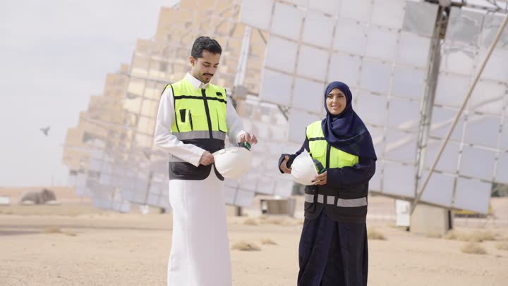 النظر الى الكاميرا بتعابير السرور و الثقة, الوقوف بأيدى متقاطعة, إرتداء خوذة وسترة الحماية, مهندسة عربية خليجية سعودية محجبة مبتسمة ترتدي العباءة السوداء تقف بجانب مهندس عربى خليجى سعودى, مهن ووظائف سعودية