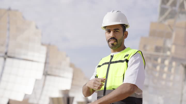 مهندس عربى خليجى سعودى يرتدى خوزة و سترة الحماية يقف بايدى متقاطعة, التدقيق والإشراف على العمل, مهن ووظائف سعودية، النظر الى الكاميرا بتعابير السرورو الثقة