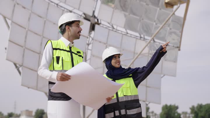 Auditing and supervising the work, pointing to something, wearing a helmet and safety vest, a smiling veiled Saudi Gulf Arab engineer wearing a black abaya standing next to a Saudi Gulf Arab engineer holding the engineering plan for the project, Saudi professions and jobs.