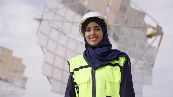 Standing with crossed arms, inspecting and supervising the work, a close-up shot of a smiling Saudi Gulf Arab female engineer wearing a black abaya, helmet, and safety vest looking at the camera with a joyful expression, Saudi professions and jobs, solar power stations.