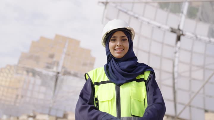 محطة الطاقة الشمسية, النظر الى الكاميرا بتعابير السرور, تصوير مقرب لمهندسة عربية خليجية سعودية محجبة مبتسمة ترتدي العباءة السوداء والخوذة وسترة الحماية, الوقوف بايدى متقاطعة, التدقيق والإشراف على العمل, مهن ووظائف سعودية،
