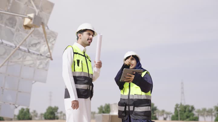الاشارة الى شىء ما, إرتداء خوذة وسترة الحماية, مهندسة عربية خليجية سعودية محجبة مبتسمة ترتدي العباءة السوداء تمسك بيدها التابلت تقف بجانب مهندس عربى خليجى سعودى يمسك مخطط هندسى, التدقيق والإشراف على العمل, مهن ووظائف سعودية