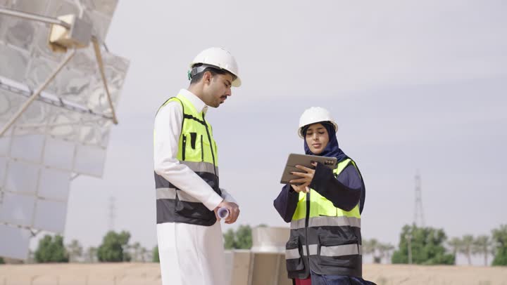 إرتداء خوذة وسترة الحماية, مهندسة عربية خليجية سعودية محجبة مبتسمة ترتدي العباءة السوداء تمسك بيدها التابلت تقف بجانب مهندس عربى خليجى سعودى يمسك مخطط هندسى, التدقيق والإشراف على العمل, مهن ووظائف سعودية،