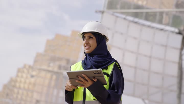 الواح وستلايت انتاج الطاقة الكهربائية, التدقيق والإشراف على العمل، تصوير مقرب لمهندسة عربية خليجية سعودية محجبة مبتسمة ترتدي العباءة السوداء و الخوذة وسترة الحماية تمسك بيدها التابلت تنظر الى شىء ما, مهن ووظائف سعودية، محطات الطاقة الشمسية