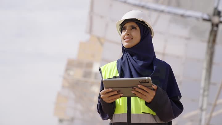 Integrating modern technology with engineering, auditing and supervising work, a close-up shot of a smiling Saudi Gulf Arab female engineer wearing a black abaya, helmet, and safety vest, holding a tablet in her hand and looking at something, Saudi professions and jobs, solar power stations.