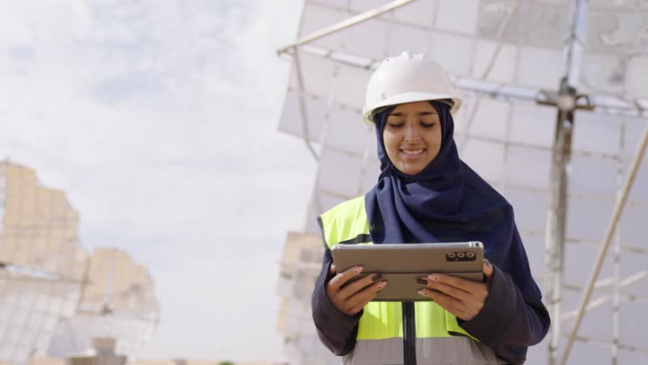 استخدام الطاقة المتجددة, التدقيق والإشراف على العمل، تصويرمقرب لمهندسة عربية خليجية سعودية محجبة مبتسمة ترتدي العباءة السوداء و الخوذة وسترة الحماية تمسك بيدها التابلت تنظر الى شىء ما, مهن ووظائف سعودية، محطات الطاقة الشمسية