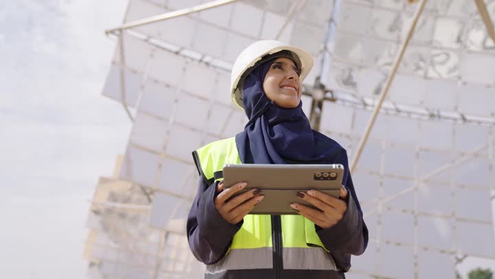 التدقيق والإشراف على العمل،  مفهوم هندسة الطاقة، تصويرمقرب لمهندسة عربية خليجية سعودية محجبة مبتسمة ترتدي الخوذة وسترة الحماية تمسك بيدها التابلت تنظر الى شىء ما, مهن ووظائف سعودية، محطات الطاقة الشمسية