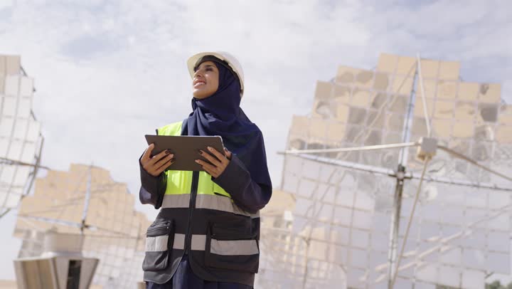 The concept of energy engineering, a close-up image of a smiling Saudi Gulf Arab engineer wearing a helmet and safety vest holding a tablet, overseeing and supervising work, Saudi professions and jobs, solar power stations.