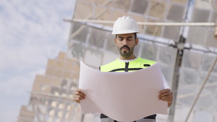 A Saudi Gulf Arab engineer wearing a helmet and safety vest holds an organizational plan in his hand, looking at something and checking the implementation of architectural engineering projects and plans in construction in Saudi Arabia, ensuring the establishment of good foundations for the building.