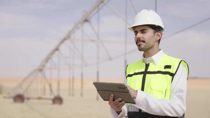 Studying the fundamentals of the project, a Saudi Gulf Arab engineer wearing traditional attire, a jacket, and a safety helmet is working on a tablet on-site, using a modern advanced technical device in the field of engineering, the concept of managing engineering and construction projects.