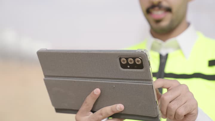 Close-up shot of a Saudi Gulf Arab engineer's hand wearing traditional attire and a safety helmet, working on a tablet at the site, using a modern advanced technical device in the field of engineering, working in the Saudi engineering sector, the concept of managing engineering and construction projects.