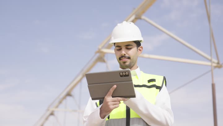 A Saudi Gulf Arab engineer wearing traditional attire, a jacket, and a safety helmet is working on a tablet at the site, using a modern advanced technical device in the field of engineering, working in the Saudi engineering sector, the concept of managing engineering and construction projects.