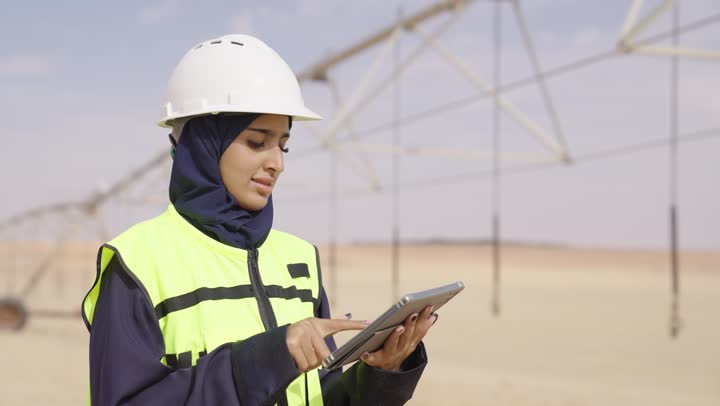A smiling Saudi Gulf Arab engineer wearing a hijab, helmet, and safety vest is using a tablet, looking at the camera, taking notes and recording observations, utilizing modern technical devices, ensuring that work is proceeding as required and correctly, and writing the latest updates on the project.