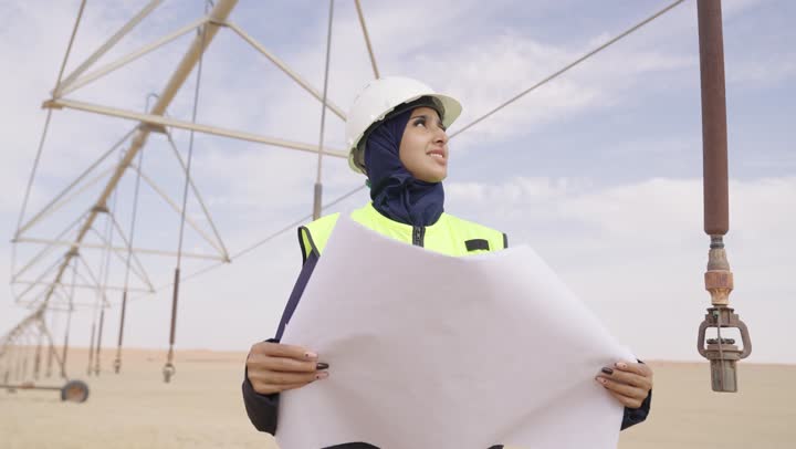 Supervising the engineering project at the work site, developing and building the Kingdom of Saudi Arabia by women, a close-up shot of a hijab-wearing Arab Gulf Saudi engineer wearing a safety helmet and vest, holding a construction plan in her hand and looking at something.
