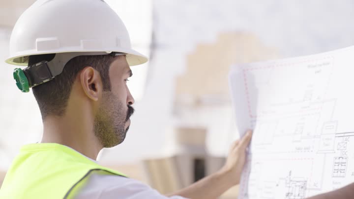 تطوير و بناء المملكة العربية السعودية على يد ابناءها, تصوير من الجانب لهندس عربي خليجي يرتدي خوذة الحماية و السترة يحمل بيده مخطط البناء، الإشراف على المشاريع الهندسية في موقع العمل