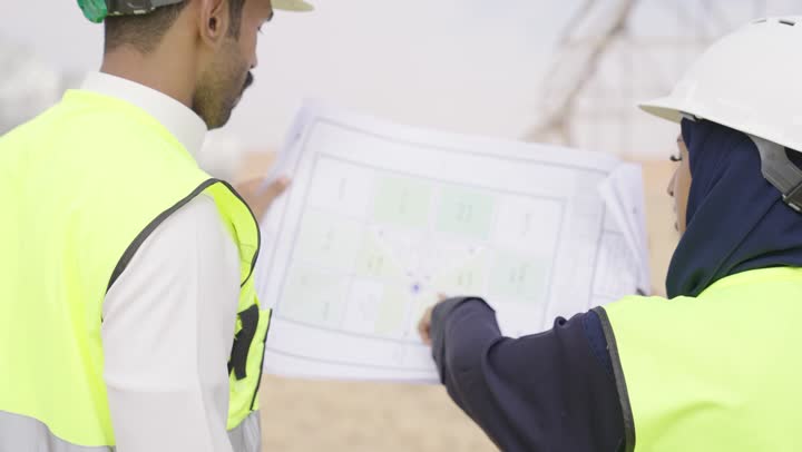 مفهوم ادارة المشاريع الهندسية، دراسة المخطط على ارض الواقع، مهنة ووظيفة هندسية، تصوير من الخلف لمهندس عربي خليجي سعودى يرتدى سترة وخوذة الحماية يحمل المخطط الهندسى و يتناقش مع زميلته حول المشروع في موقع البناء