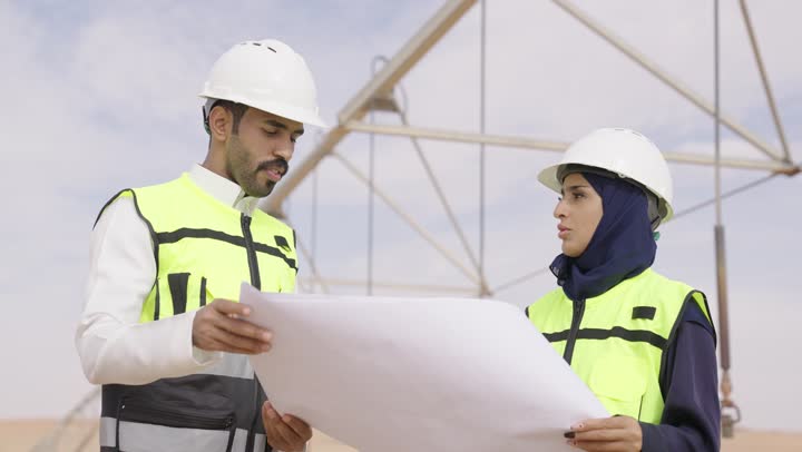 وظيفة ومهنة هندسية، دراسة المخطط على ارض الواقع مهندس عربي خليجي سعودى يرتدي خوذة و سترة الحماية يعمل مع زميلته في القطاع الهندسي في السعودية، ادارة المشاريع الهندسية والانشاءات باستخدام التكنولوجيا