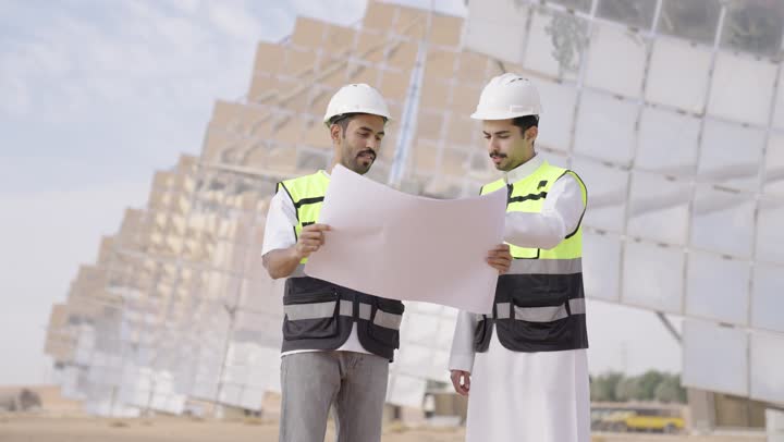 Auditing and supervising the work, two Arab Gulf engineers from Saudi Arabia wearing helmets and safety vests holding a plan and engineering design in their hands, the concept of energy engineering, Saudi professions and jobs, the future of energy, professional energy engineer.