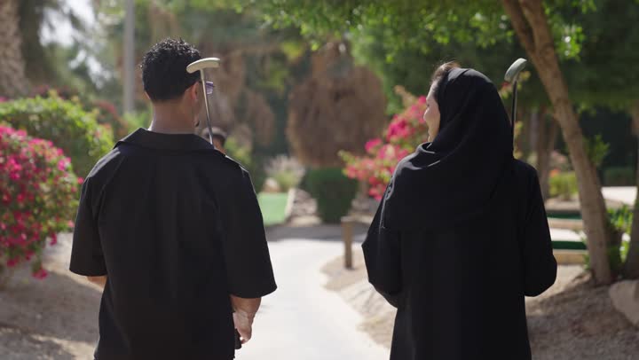 أنشطة رياضية بالسعودية، تصوير من الخلف لشاب عربي خليجي سعودي يرتدي الزي الكاجوال يحمل عصا اللعب يمشي برفقة زوجته، ممارسة لعبة الغولف على مساحات واسعة خضراء، رياضات الألعاب الأولمبية الصيفية